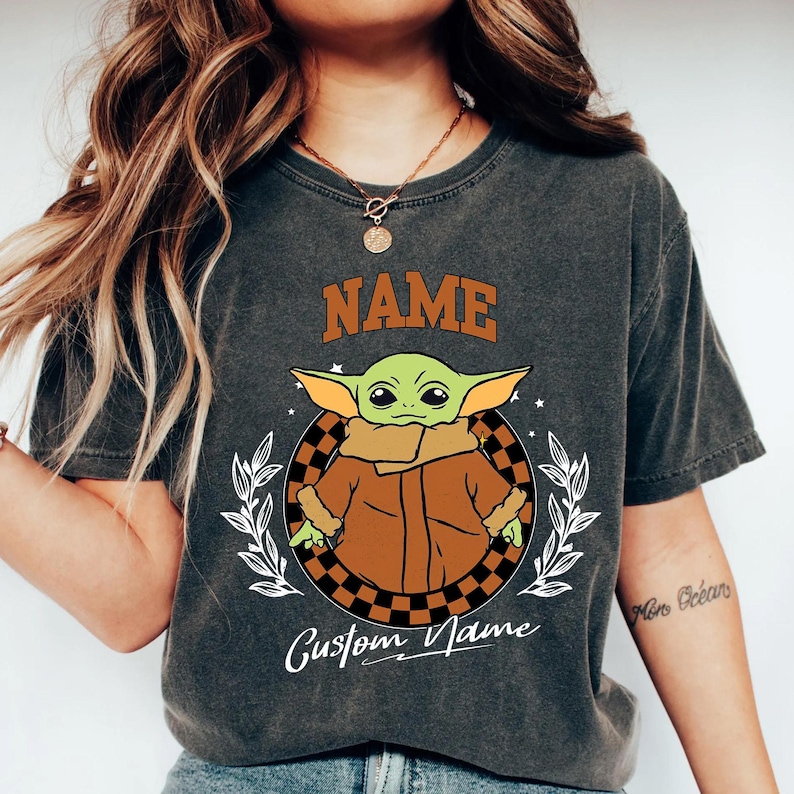 K&ouml;nnte beinhalten: Dunkelgraues T-Shirt mit einer Cartoonfigur in einem braunen Mantel, die sich in einem karierten Kreis befindet. Das Wort "NAME" steht &uuml;ber der Figur, und "Custom Name" steht darunter. Wei&szlig;e Bl&auml;tter und Sterne umgeben den Kreis.