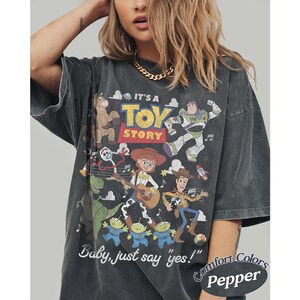 May include: A charcoal gray short-sleeve t-shirt featuring a graphic print of Toy Story characters including Woody, Jessie, Buzz Lightyear, Rex, Bullseye, and Forky. The text reads IT'S A TOY STORY and Baby, just say yes! with Comfort Colors Pepper.