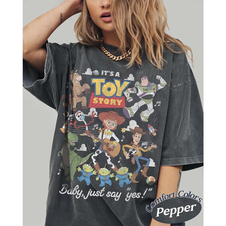 May include: A charcoal gray short-sleeve t-shirt featuring a graphic print of Toy Story characters including Woody, Jessie, Buzz Lightyear, Rex, Bullseye, and Forky. The text reads IT'S A TOY STORY and Baby, just say yes! with Comfort Colors Pepper.
