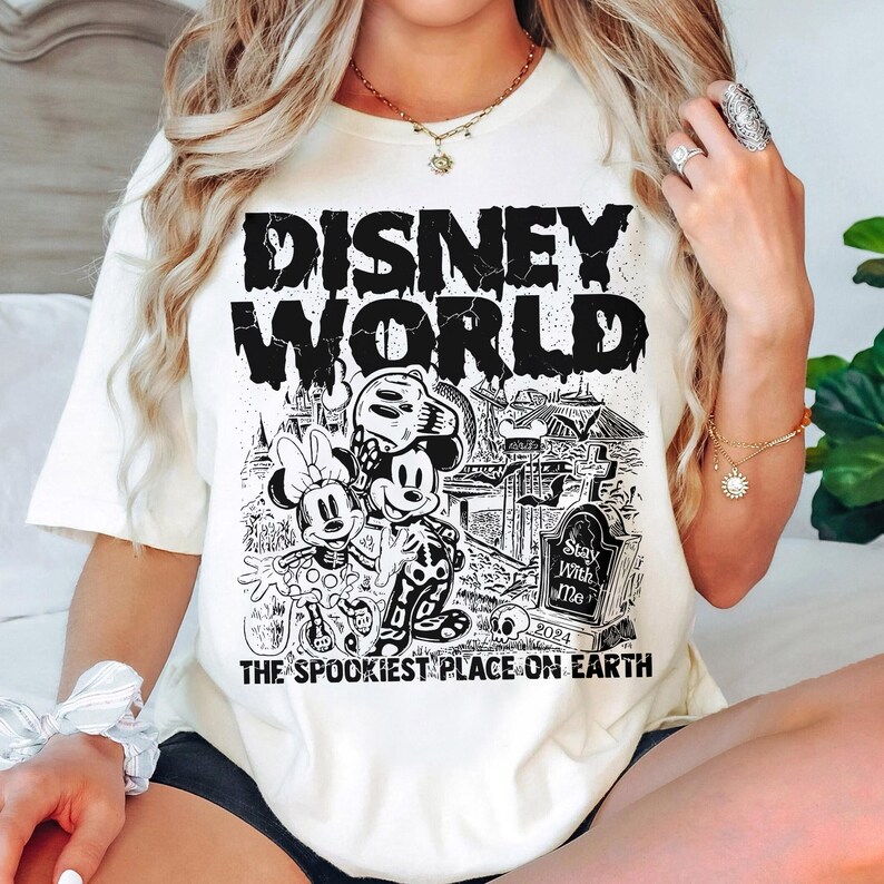 K&ouml;nnte beinhalten: Ein wei&szlig;es T-Shirt mit einem schwarzen Grafikdruck von Mickey Mouse und Minnie Mouse, die als Skelette verkleidet sind. Die Grafik enth&auml;lt auch den Text "Disney World", "The Spookiest Place on Earth" und "Stay With Me 2024".