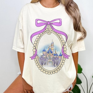 May include: A white t-shirt with a purple bow and a pearl-encrusted frame surrounding a cartoon image of a castle. The castle is in a light blue, pink, and yellow color scheme.