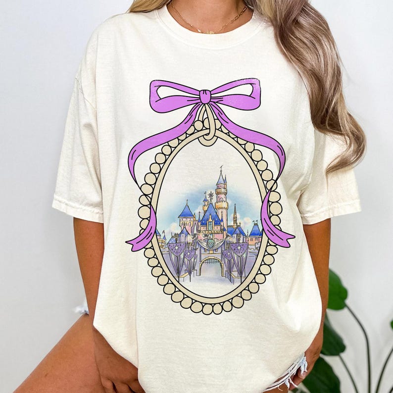 May include: A white t-shirt with a purple bow and a pearl-encrusted frame surrounding a cartoon image of a castle. The castle is in a light blue, pink, and yellow color scheme.