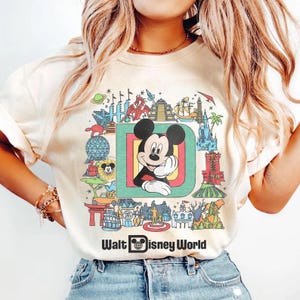 Puede incluir: Camiseta color crema con un gr&aacute;fico colorido de Mickey Mouse dentro de una gran letra "D" y varios monumentos de Disney World. Las palabras "Walt Disney World" est&aacute;n impresas debajo.