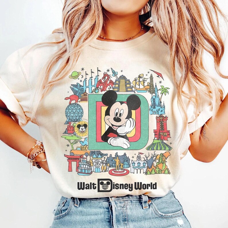 Puede incluir: Camiseta color crema con un gr&aacute;fico colorido de Mickey Mouse dentro de una gran letra "D" y varios monumentos de Disney World. Las palabras "Walt Disney World" est&aacute;n impresas debajo.