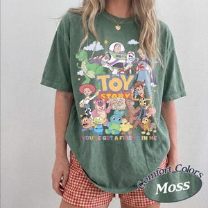 May include: A moss green short-sleeve t-shirt featuring a colorful graphic print of various Toy Story characters. The text Toy Story and You've Got A Friend In Me are printed on the front. The shirt has a crew neckline and a relaxed fit.