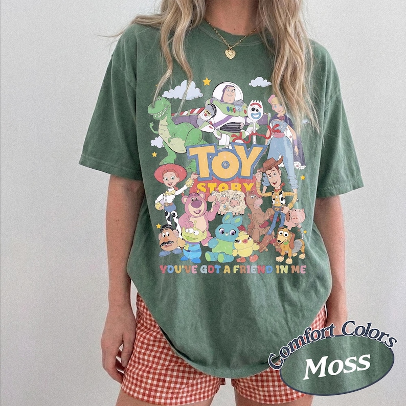May include: A moss green short-sleeve t-shirt featuring a colorful graphic print of various Toy Story characters. The text Toy Story and You've Got A Friend In Me are printed on the front. The shirt has a crew neckline and a relaxed fit.