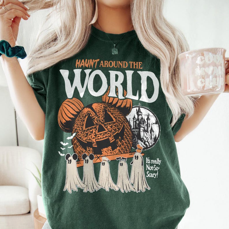 May include: Green t-shirt with a vintage-style graphic of a pumpkin-faced Mickey Mouse head, a castle, and ghosts. The text "HAUNT AROUND THE WORLD" is printed above the graphic. The text "It's really Not-So-Scary!" is printed below the graphic.