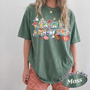 May include: A moss green short-sleeve t-shirt featuring a graphic print with the text I KNEW A I KNEW YOU and various cartoon characters including a cowboy, a space ranger, a pig, and a horse, along with a red heart shape containing the letters TS.