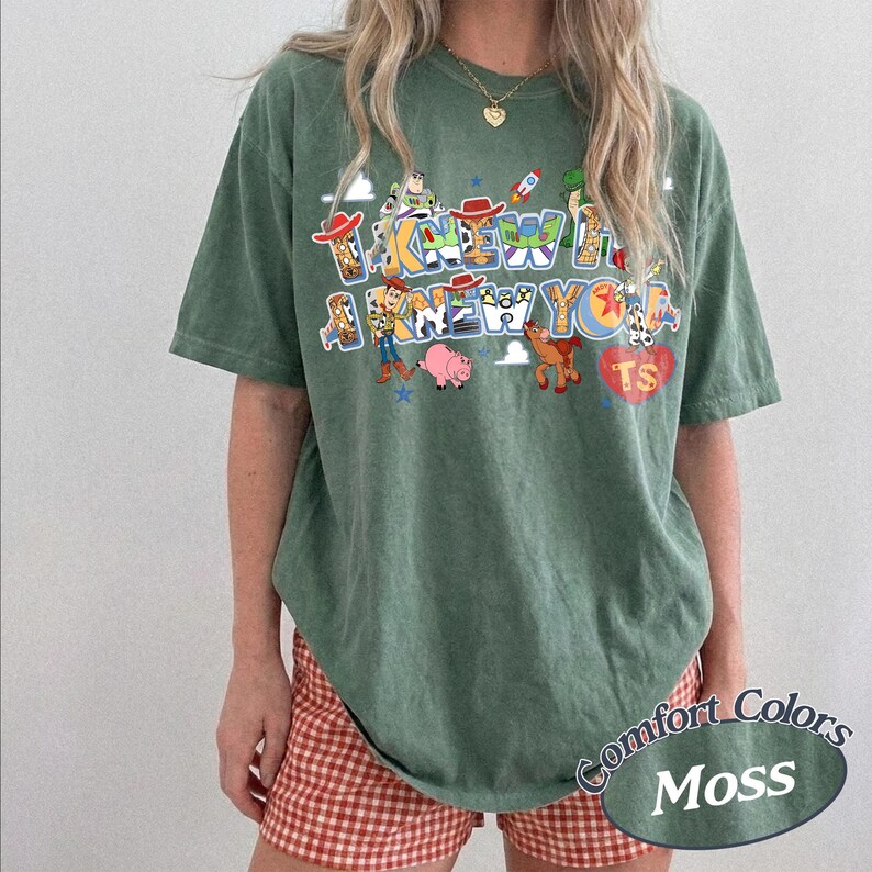 May include: A moss green short-sleeve t-shirt featuring a graphic print with the text I KNEW A I KNEW YOU and various cartoon characters including a cowboy, a space ranger, a pig, and a horse, along with a red heart shape containing the letters TS.