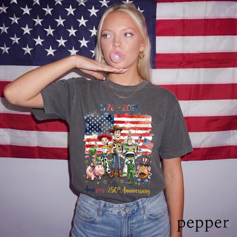 May include: A charcoal grey t-shirt featuring a graphic of Toy Story characters including Woody, Buzz Lightyear, Jessie, Rex, Hamm, Mr. Potato Head, and a green army soldier. The text reads 1776-2026 and America 250th Anniversary with fireworks.