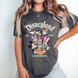 May include: Dark gray t-shirt featuring the Disneyland logo with Mickey Mouse, Minnie Mouse, Donald Duck, Daisy Duck, Goofy, and Pluto. The shirt also includes the text "Est 1955" and "California".