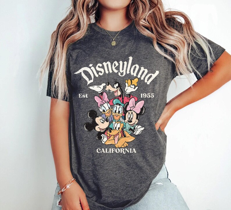 May include: Dark gray t-shirt featuring the Disneyland logo with Mickey Mouse, Minnie Mouse, Donald Duck, Daisy Duck, Goofy, and Pluto. The shirt also includes the text "Est 1955" and "California".