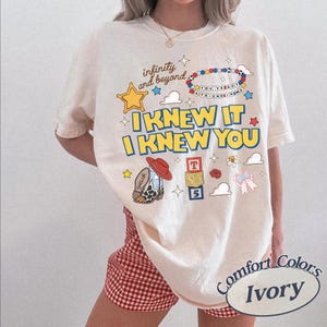 May include: An ivory t-shirt featuring a graphic print with the text "infinity and beyond", "YOU'VE GOT A FRIEND IN ME", and "I KNEW IT I KNEW YOU". The design includes a yellow star, a beaded bracelet, a cowboy boot, alphabet blocks, and clouds.