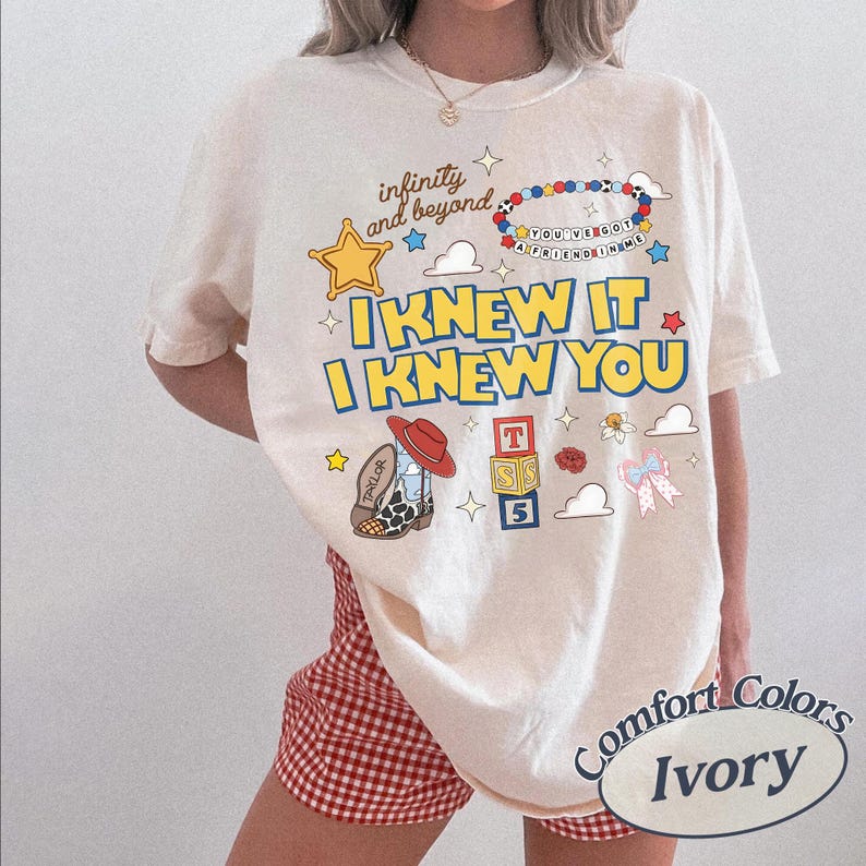 May include: An ivory t-shirt featuring a graphic print with the text "infinity and beyond", "YOU'VE GOT A FRIEND IN ME", and "I KNEW IT I KNEW YOU". The design includes a yellow star, a beaded bracelet, a cowboy boot, alphabet blocks, and clouds.