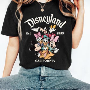 May include: Black t-shirt featuring a graphic of Disneyland characters including Mickey Mouse, Minnie Mouse, Donald Duck, and Goofy. The text "Disneyland" is arched across the top, with "Est 1955" below. "CALIFORNIA" is printed at the bottom.