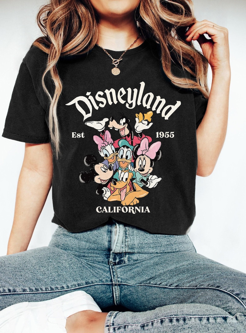 May include: Black t-shirt featuring a graphic of Disneyland characters including Mickey Mouse, Minnie Mouse, Donald Duck, and Goofy. The text "Disneyland" is arched across the top, with "Est 1955" below. "CALIFORNIA" is printed at the bottom.