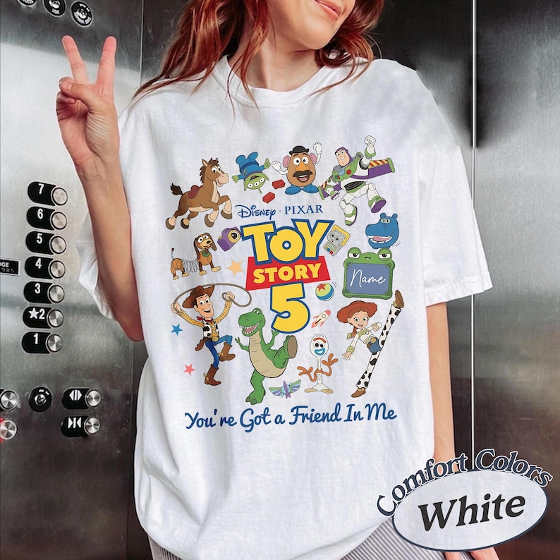 May include: A white t-shirt featuring a colorful graphic of Toy Story characters including Woody, Buzz Lightyear, Rex, and Slinky Dog. The design includes the text Disney Pixar Toy Story 5 and You've Got a Friend In Me, with a blank space for a name.