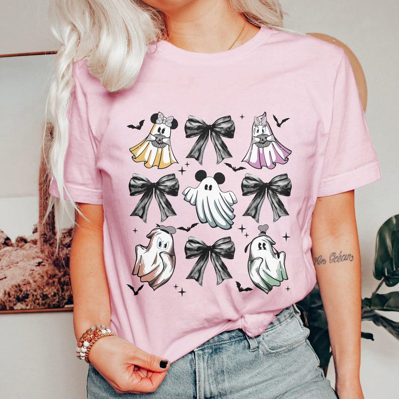 May include: A light pink t-shirt featuring a Halloween-themed design. The design includes cartoon ghosts with black bows, bats, and cross symbols. The ghosts are in various colors, including yellow, white, and purple.