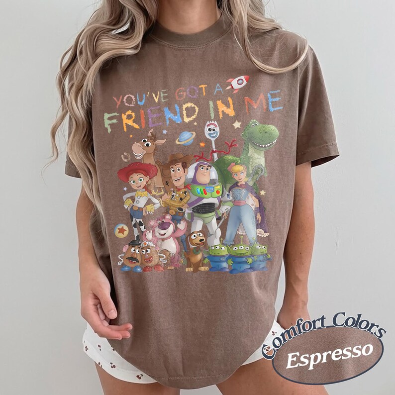 May include: A brown short-sleeve t-shirt featuring a colorful graphic print of various animated characters from the Toy Story franchise. The text "YOU'VE GOT A FRIEND IN ME" is printed in a multi-colored, distressed font above the character group.