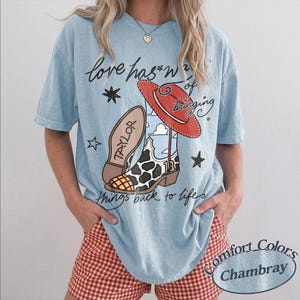 May include: A light blue t-shirt featuring a graphic print of a cow-print cowboy boot with the number 13, a red cowboy hat, and stars. The text reads, "love has a way of bringing things back to life" with the name "TAYLOR" on the boot sole.