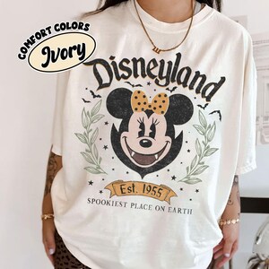 May include: Ivory-colored t-shirt with a Disneyland graphic featuring Minnie Mouse with a polka-dot bow. The design includes bats, stars, and the text "Est. 1955 Spookiest Place on Earth."