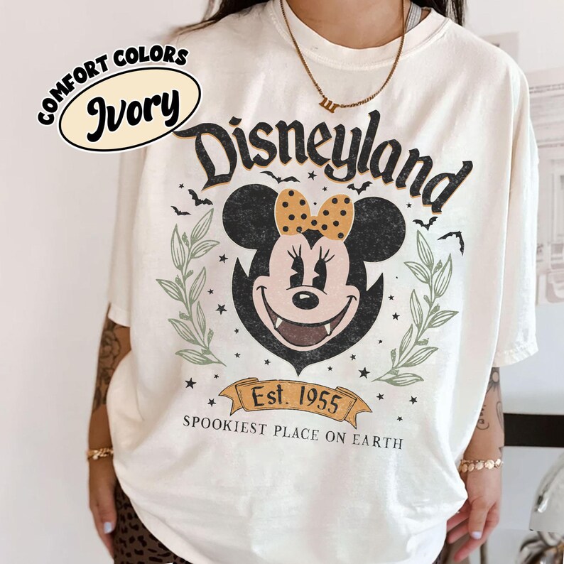 May include: Ivory-colored t-shirt with a Disneyland graphic featuring Minnie Mouse with a polka-dot bow. The design includes bats, stars, and the text "Est. 1955 Spookiest Place on Earth."