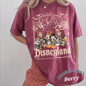 May include: Berry-colored t-shirt with a Disneyland graphic featuring Disney characters in skeleton costumes. The graphic includes a haunted house, bats, and the text "Est. 1955". The shirt is a Comfort Colors brand.