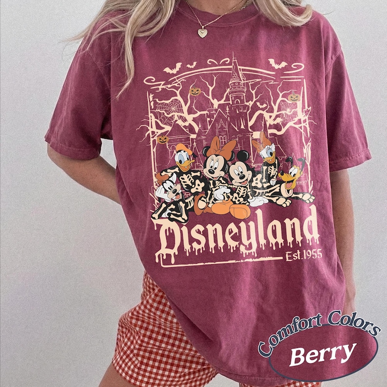 May include: Berry-colored t-shirt with a Disneyland graphic featuring Disney characters in skeleton costumes. The graphic includes a haunted house, bats, and the text "Est. 1955". The shirt is a Comfort Colors brand.