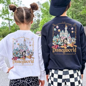 May include: Two children wearing white and black sweatshirts with a graphic of Mickey Mouse and friends in front of Cinderella's Castle. The text "Disneyworld -1971- " is printed on the sweatshirts.