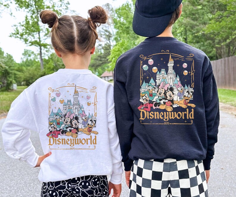 May include: Two children wearing white and black sweatshirts with a graphic of Mickey Mouse and friends in front of Cinderella's Castle. The text "Disneyworld -1971- " is printed on the sweatshirts.
