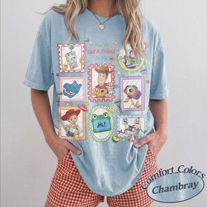 May include: A light blue t-shirt featuring a graphic print of various cartoon characters arranged in postage stamp-style frames. The text "You've Got A Friend In Me" is printed in the center, along with a small green frog graphic labeled "Hi!"