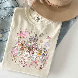 May include: Cream-colored t-shirt featuring a Disney World graphic with Mickey Mouse, Minnie Mouse, Donald Duck, and other characters. The design includes a castle, balloons, and the words "Disneyworld" in pastel colors. A tan hat and blue jeans are in the background.