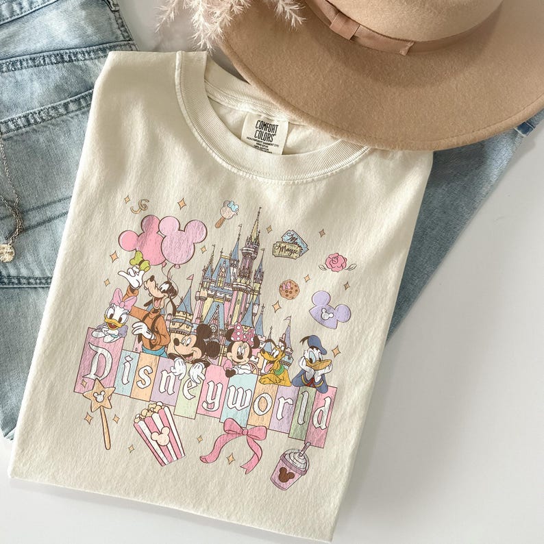 May include: Cream-colored t-shirt featuring a Disney World graphic with Mickey Mouse, Minnie Mouse, Donald Duck, and other characters. The design includes a castle, balloons, and the words "Disneyworld" in pastel colors. A tan hat and blue jeans are in the background.