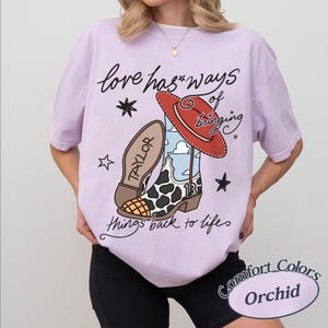 May include: A light purple t-shirt featuring a graphic of a cowboy boot with a cow print pattern, a red hat, and the text "love has ways of bringing things back to life" along with the name "TAYLOR" and the number "13" on the boot design.
