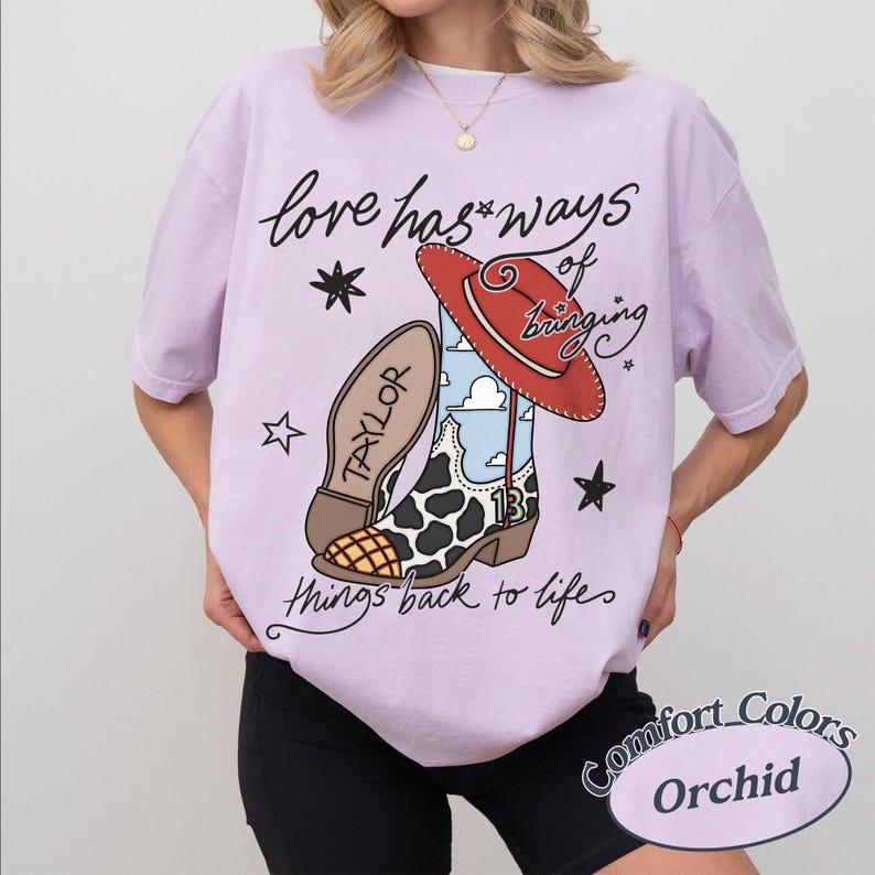 May include: A light purple t-shirt featuring a graphic of a cowboy boot with a cow print pattern, a red hat, and the text "love has ways of bringing things back to life" along with the name "TAYLOR" and the number "13" on the boot design.