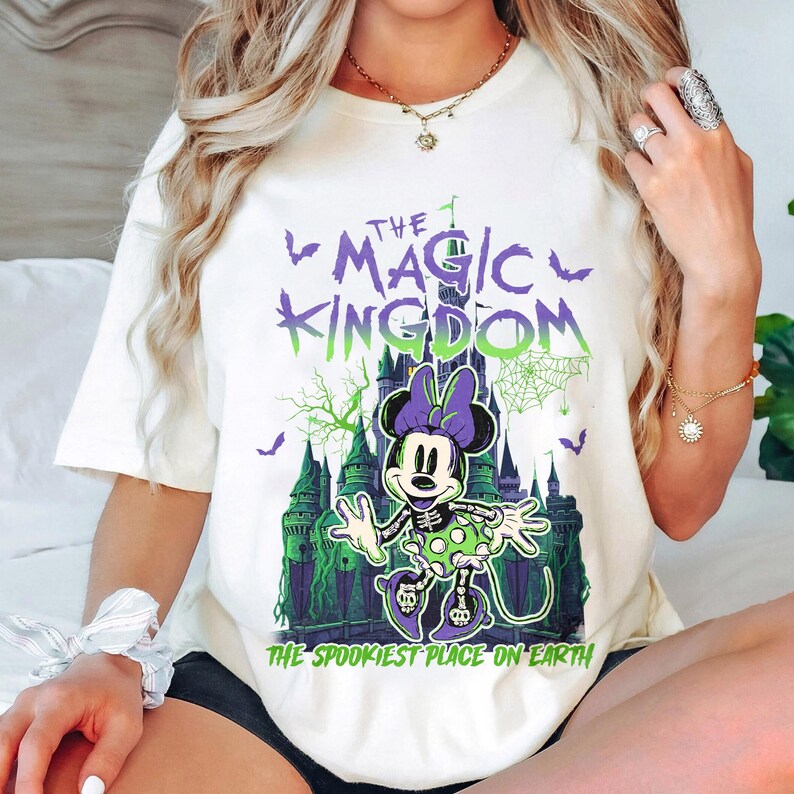 May include: Cream-colored t-shirt featuring a Halloween-themed graphic. The design includes the text "The Magic Kingdom" and "The Spookiest Place on Earth," along with a cartoon Minnie Mouse in a skeleton costume in front of a castle.