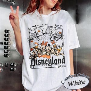 May include: White t-shirt featuring a Halloween-themed Disneyland graphic. The design includes Mickey and Minnie Mouse, Donald and Daisy Duck, and Goofy in skeleton costumes, with a haunted castle and the text "Disneyland Est.1955".
