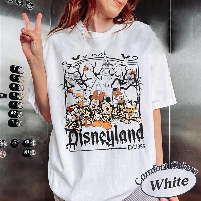 May include: White t-shirt featuring a Halloween-themed Disneyland graphic. The design includes Mickey and Minnie Mouse, Donald and Daisy Duck, and Goofy in skeleton costumes, with a haunted castle and the text "Disneyland Est.1955".