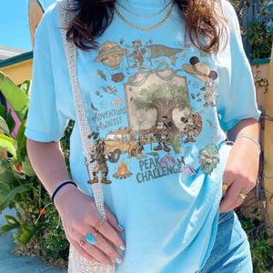 May include: Light blue t-shirt with a colorful graphic featuring cartoon characters, a tree, and the text "Adventure Awaits!" and "Peak Challenge". The design includes a jeep, hot air balloon, and a campfire.