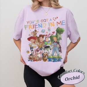 May include: A light purple short-sleeve t-shirt featuring a colorful graphic of various Toy Story characters and the text "YOU'VE GOT A FRIEND IN ME" printed across the chest. The design includes Woody, Buzz Lightyear, Jessie, and others.