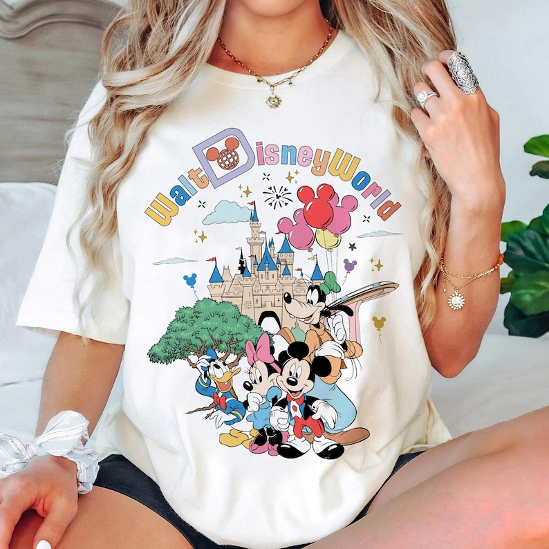 May include: White t-shirt with a colorful graphic of Mickey Mouse, Minnie Mouse, Goofy, Donald Duck, and the Disney castle. The text "Walt Disney World" is printed in a rainbow arc around the graphic.