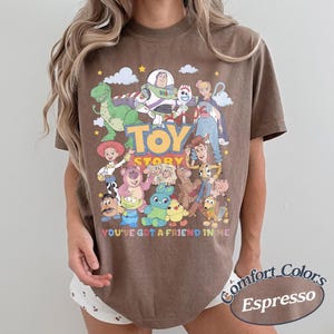 May include: A brown short-sleeve t-shirt featuring a graphic print of Toy Story characters including Woody, Buzz Lightyear, Jessie, and Rex. The text Toy Story and You've Got A Friend In Me are printed in colorful letters across the front of the shirt.