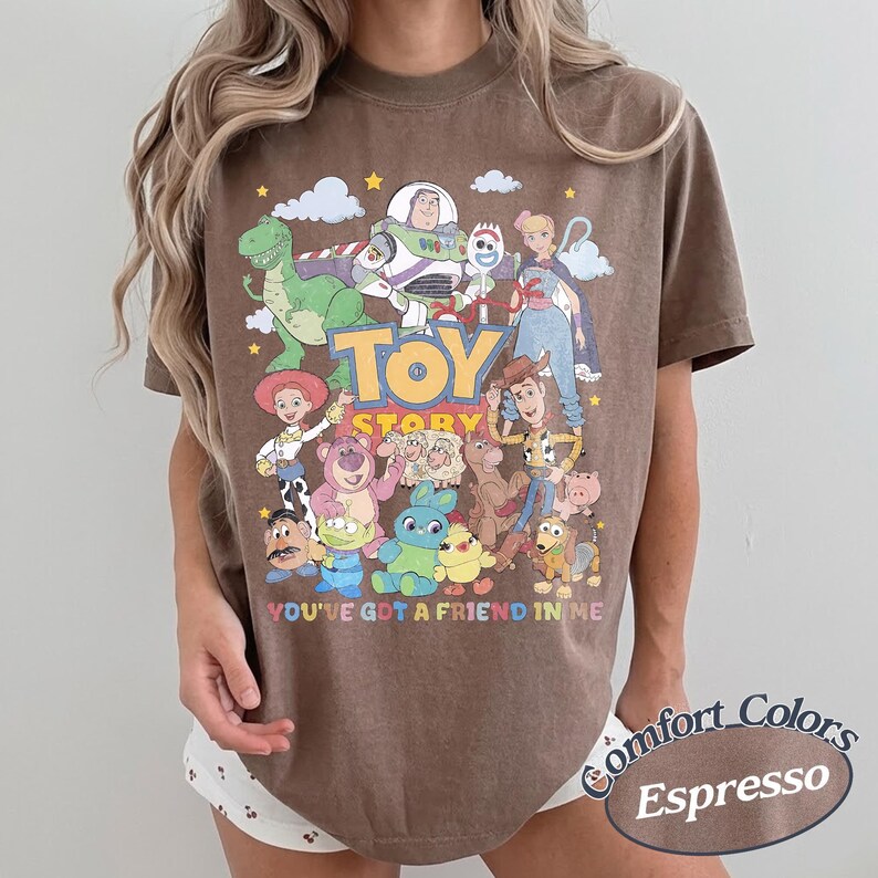 May include: A brown short-sleeve t-shirt featuring a graphic print of Toy Story characters including Woody, Buzz Lightyear, Jessie, and Rex. The text Toy Story and You've Got A Friend In Me are printed in colorful letters across the front of the shirt.