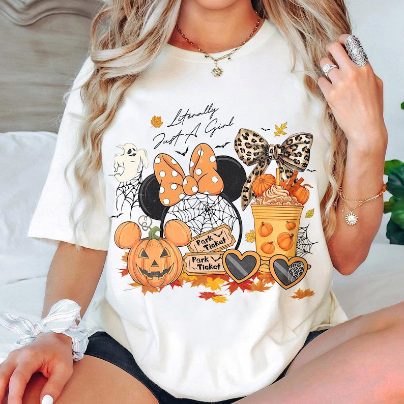 May include: Cream-colored t-shirt with a Halloween-themed graphic. The design includes a Minnie Mouse head with an orange polka dot bow, a pumpkin, and the text "Literally Just A Girl". Other elements include a ghost, spiderwebs, and fall leaves.