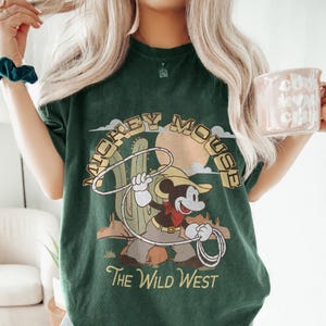 May include: Dark green t-shirt with a vintage Mickey Mouse cowboy graphic, holding a lasso against a desert backdrop. The text "MICKEY MOUSE" is arched above the image, with "The Wild West" below.