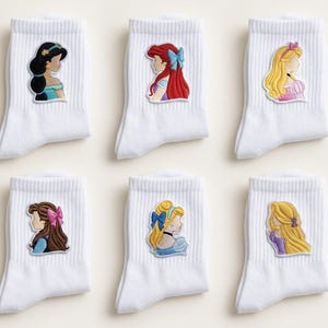 May include: Six pairs of white ankle socks, each with a princess design. The embroidered patches show the princesses in profile, with their iconic hairstyles and outfits. The socks are made of a ribbed knit material.