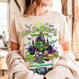 May include: Beige t-shirt with a vibrant graphic promoting Mickey's Not-So-Scary Halloween Party at Walt Disney World. The design showcases Mickey, Minnie, Donald, Goofy, and Pluto in Halloween attire, set against a spooky castle scene.