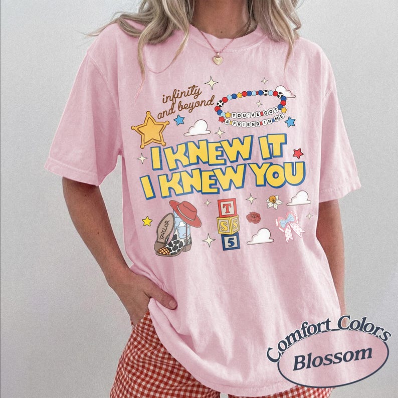 May include: A light pink short-sleeve t-shirt featuring a graphic print with the text "infinity and beyond", "I KNEW IT I KNEW YOU", and "YOU'VE GOT A FRIEND IN ME". The design includes a yellow star, a beaded bracelet, a cowboy boot, and alphabet blocks.