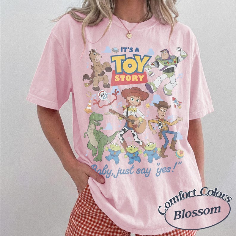 May include: A light pink short-sleeve t-shirt featuring a graphic print of Toy Story characters including Woody, Buzz Lightyear, Jessie, Bullseye, Rex, Forky, and the Aliens. The text reads IT'S A TOY STORY and Baby, just say yes! in blue and red.