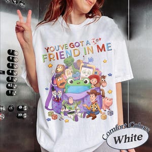 May include: A white t-shirt featuring a colorful graphic of various animated characters from the Toy Story franchise emerging from a purple backpack, with the text "YOU'VE GOT A FRIEND IN ME" printed in multicolored letters above the illustration.